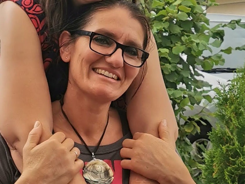 Zwei Frauen sitzen eng beieinander im Freien. Die Frau im Hintergrund umarmt die Frau im Vordergrund von hinten über die Schultern. Beide tragen Brillen und lächeln. Im Vordergrund steht ein weißer Becher auf einem Tisch. Im Hintergrund sind grüne Pflanzen und Bäume zu sehen. Zwei Frauen sitzen eng beieinander im Freien. Die Frau im Hintergrund umarmt die Frau im Vordergrund von hinten über die Schultern. Beide tragen Brillen und lächeln. Im Vordergrund steht ein weißer Becher auf einem Tisch. Im Hintergrund sind grüne Pflanzen und Bäume zu sehen.