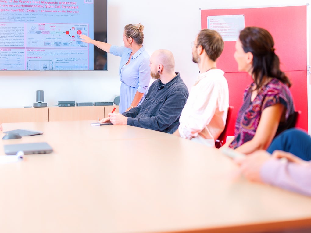 Eine Frau, die auf einen Bildschirm zeigt in einen Raum voller Personen Eine Frau, die auf einen Bildschirm zeigt in einen Raum voller Personen