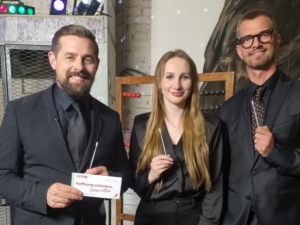 Zwei Männer und eine Frau stehen nebeneinander und blicken lächelnd in die Kamera. Alle drei tragen dunkle Kleidung. Die Frau und der Mann rechts halten jeweils ein Wattestäbchen in der Hand. Der Mann links hält zusätzlich ein DKMS-Kärtchen mit der Aufschrift „Mund auf. Stäbchen rein. Spender sein.“. Im Hintergrund sind bunte Lichter und metallische Regale zu sehen. Zwei Männer und eine Frau stehen nebeneinander und blicken lächelnd in die Kamera. Alle drei tragen dunkle Kleidung. Die Frau und der Mann rechts halten jeweils ein Wattestäbchen in der Hand. Der Mann links hält zusätzlich ein DKMS-Kärtchen mit der Aufschrift „Mund auf. Stäbchen rein. Spender sein.“. Im Hintergrund sind bunte Lichter und metallische Regale zu sehen.