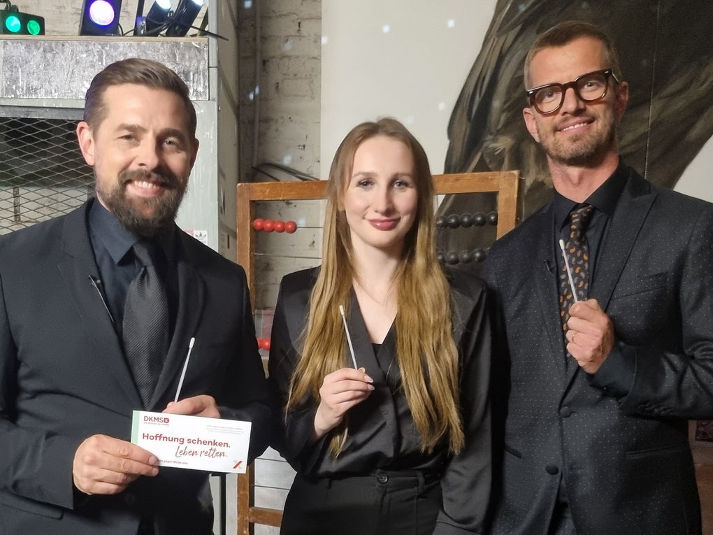 Zwei Männer und eine Frau stehen nebeneinander und blicken lächelnd in die Kamera. Alle drei tragen dunkle Kleidung. Die Frau und der Mann rechts halten jeweils ein Wattestäbchen in der Hand. Der Mann links hält zusätzlich ein DKMS-Kärtchen mit der Aufschrift „Mund auf. Stäbchen rein. Spender sein.“. Im Hintergrund sind bunte Lichter und metallische Regale zu sehen. Zwei Männer und eine Frau stehen nebeneinander und blicken lächelnd in die Kamera. Alle drei tragen dunkle Kleidung. Die Frau und der Mann rechts halten jeweils ein Wattestäbchen in der Hand. Der Mann links hält zusätzlich ein DKMS-Kärtchen mit der Aufschrift „Mund auf. Stäbchen rein. Spender sein.“. Im Hintergrund sind bunte Lichter und metallische Regale zu sehen.