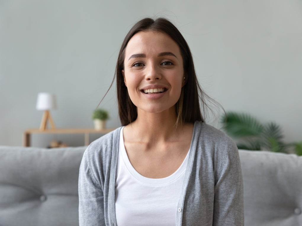 Junge Frau mit langen dunklen Haaren sitzt lächelnd auf einem grauen Sofa. Sie trägt ein weißes Shirt und eine graue Strickjacke. Im Hintergrund sind eine kleine Lampe, eine Pflanze und ein Regal zu sehen. Junge Frau mit langen dunklen Haaren sitzt lächelnd auf einem grauen Sofa. Sie trägt ein weißes Shirt und eine graue Strickjacke. Im Hintergrund sind eine kleine Lampe, eine Pflanze und ein Regal zu sehen.