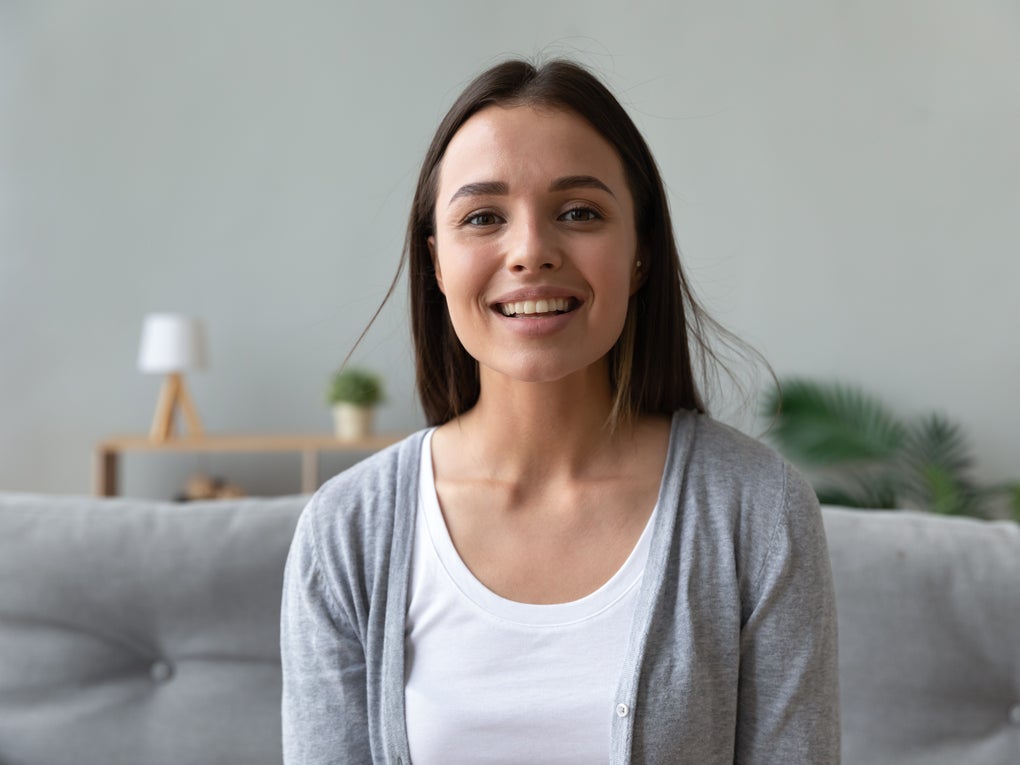 Junge Frau mit langen dunklen Haaren sitzt lächelnd auf einem grauen Sofa. Sie trägt ein weißes Shirt und eine graue Strickjacke. Im Hintergrund sind eine kleine Lampe, eine Pflanze und ein Regal zu sehen. Junge Frau mit langen dunklen Haaren sitzt lächelnd auf einem grauen Sofa. Sie trägt ein weißes Shirt und eine graue Strickjacke. Im Hintergrund sind eine kleine Lampe, eine Pflanze und ein Regal zu sehen.