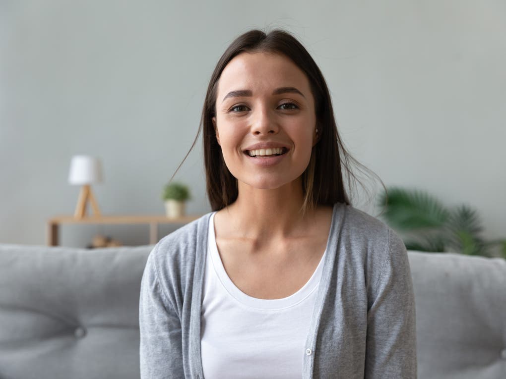 Junge Frau mit langen dunklen Haaren sitzt lächelnd auf einem grauen Sofa. Sie trägt ein weißes Shirt und eine graue Strickjacke. Im Hintergrund sind eine kleine Lampe, eine Pflanze und ein Regal zu sehen. Junge Frau mit langen dunklen Haaren sitzt lächelnd auf einem grauen Sofa. Sie trägt ein weißes Shirt und eine graue Strickjacke. Im Hintergrund sind eine kleine Lampe, eine Pflanze und ein Regal zu sehen.