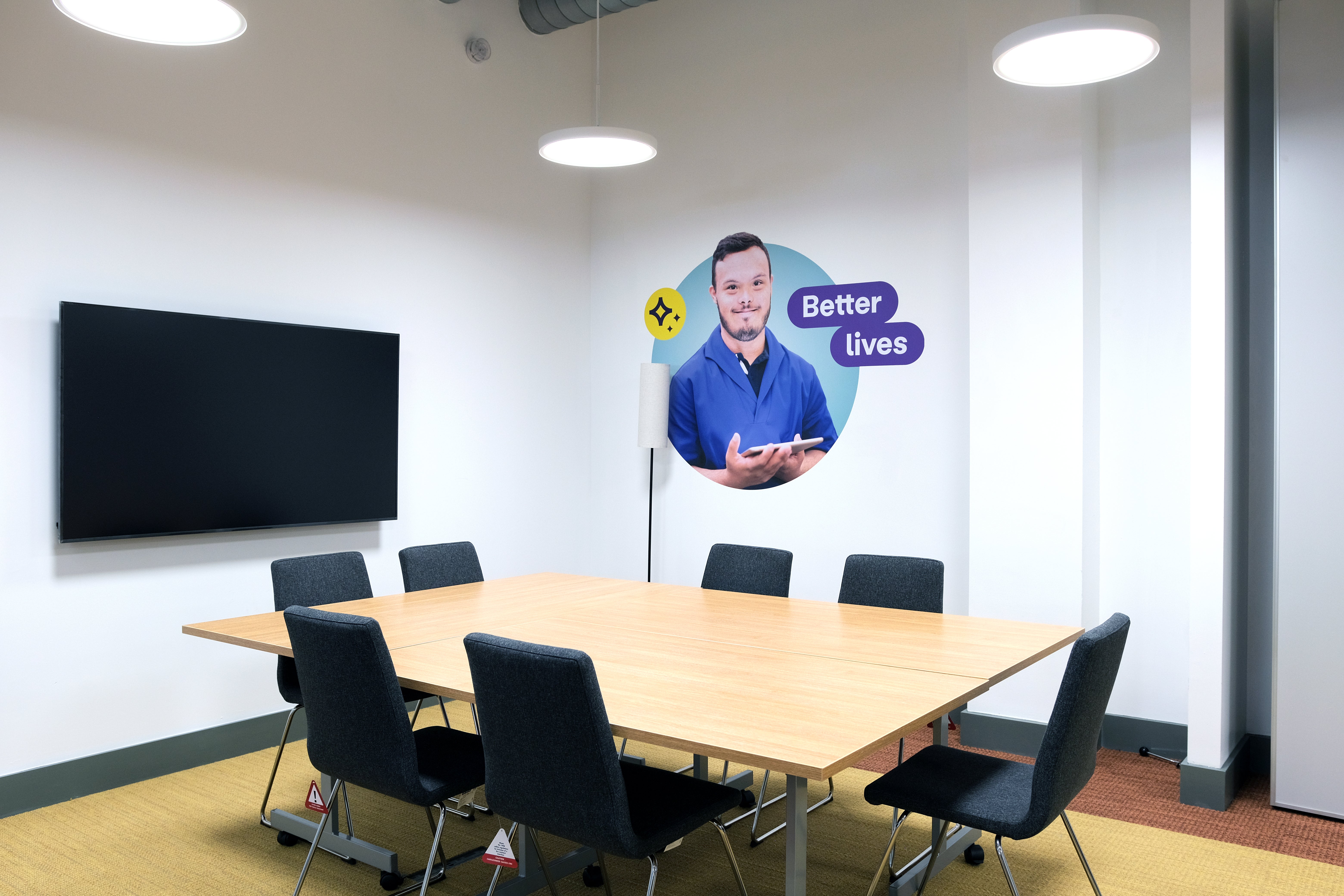 Meeting Room 1 with a table, 7 chairs, one monitor and an image on the wall with a man and a quote saying "Better lives".