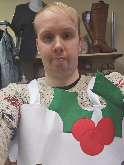 A selfie of Tom. Tom has blonde hair and is wearing a cosy jumper and a Christmas apron with some holly on it.