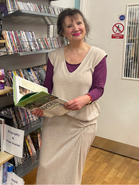 Ruth is standing in the book section of the shop. She is holding a book and smiling at the camera.