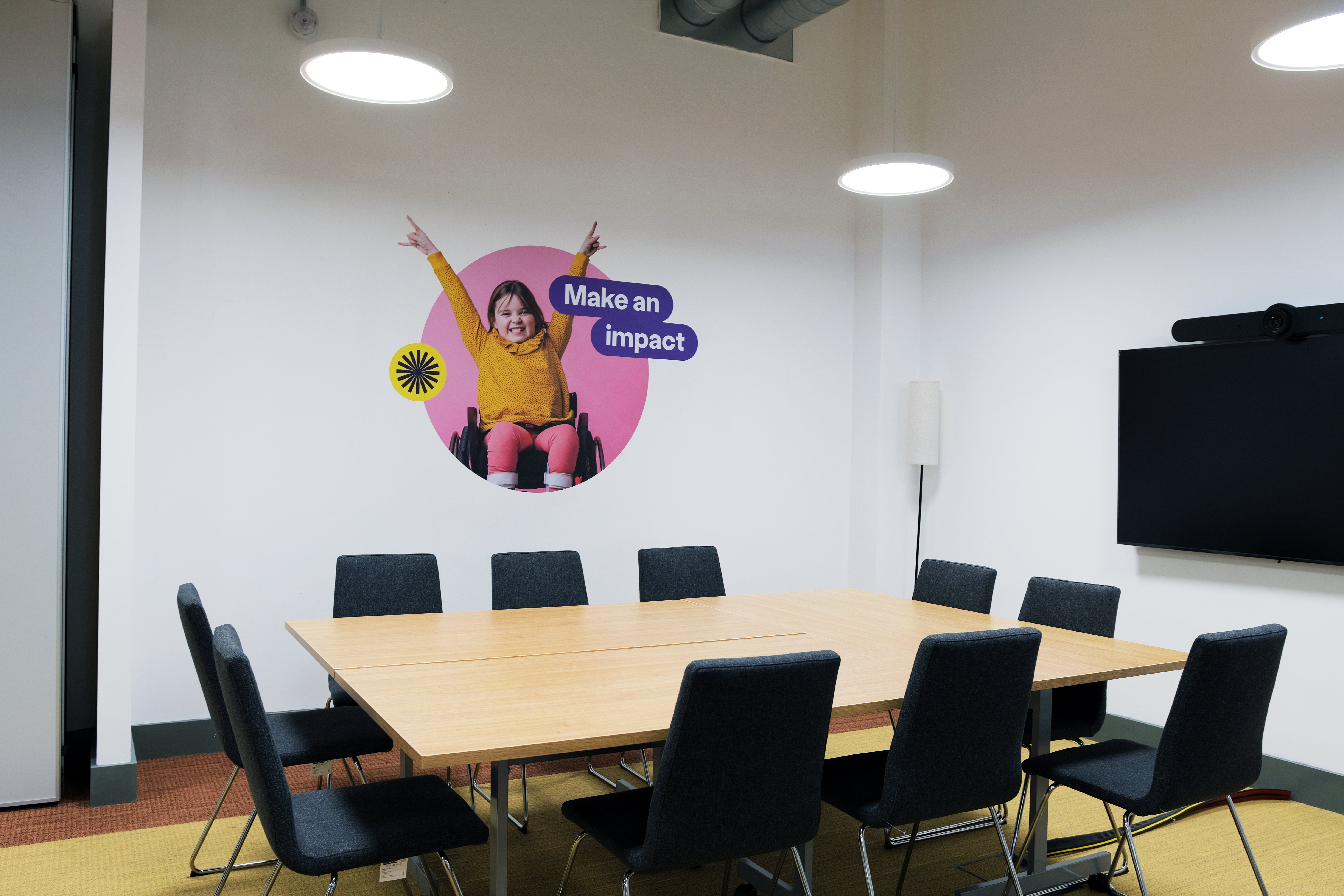 A table with 10 chairs, one conference system and image on the wall with a girl on a wheelchair saying "Make an impact".