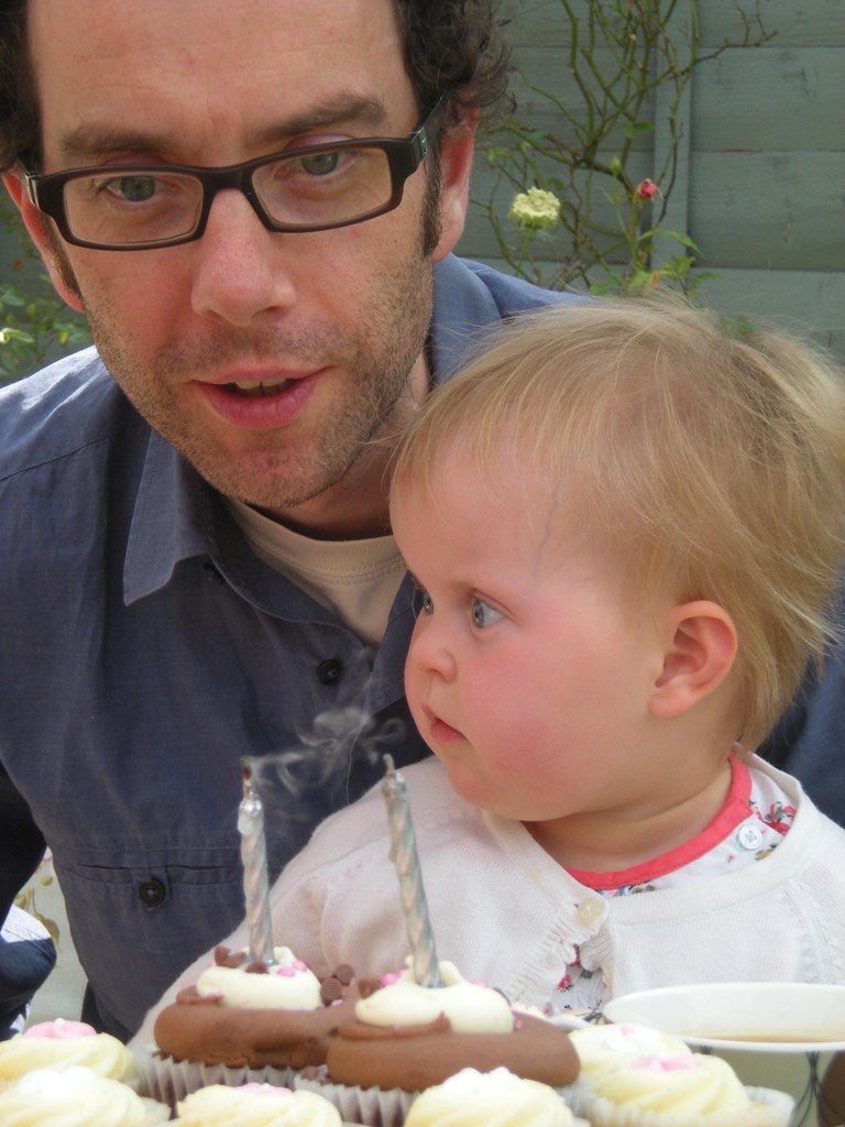 A man in glasses smiles with his baby. The baby sits in front of cupcakes with birthday candles in.