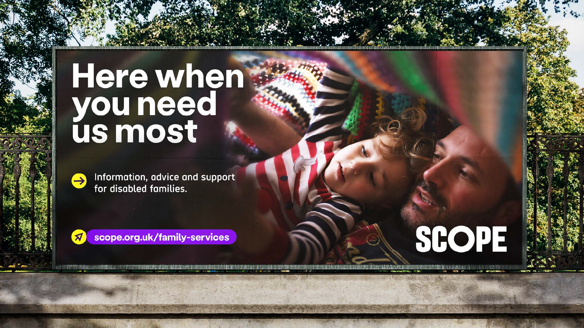 Billboard with reportage photography of a man reading a book to his child under a crochet blanket. To the left, text says 'Here when you need us most' then our arrow icon pointing to text 'Information, advice and support for disabled families'. Below this a mouse point icon and purple lozenge with scope.org.uk/family-services. Our logo is in bottom right.