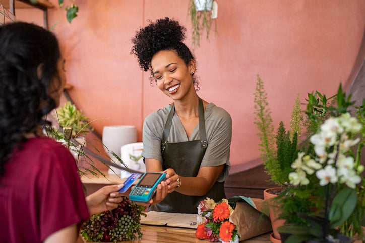 Smiling and friendly florist holding card reader machine at counter with customer paying with credit card.