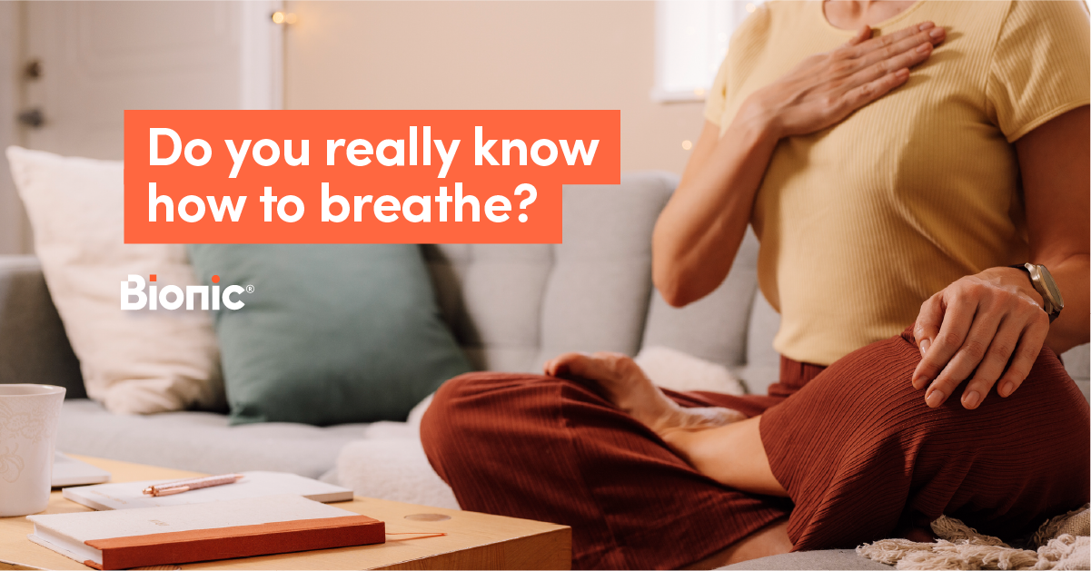 Woman in brown trousers and beige shirt sits cross-legged on sofa with her hand on her chest doing breathing exercises