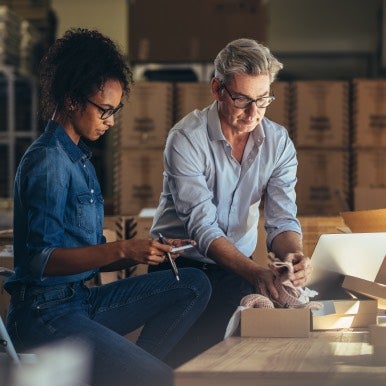 Man packing small item into box while woman looks on with calculator and pen