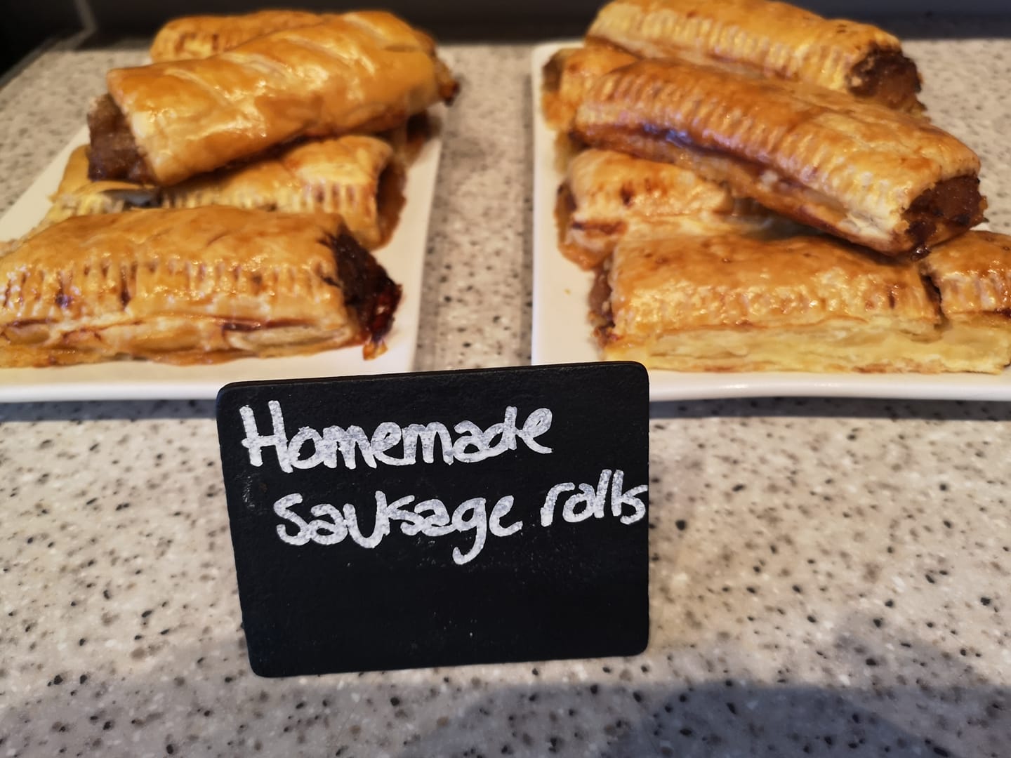 Two plates of sausage rolls on a counter behind a black sign reading 'homemade sausage rolls'