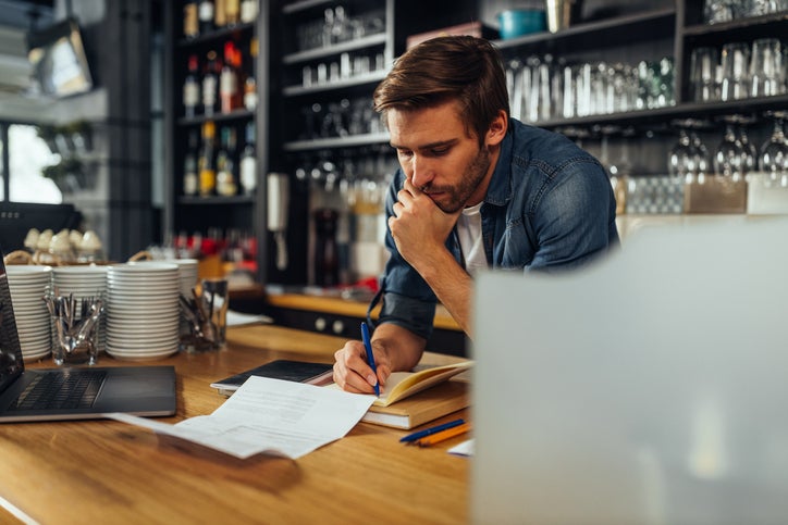 A business owner stands behind the counter at his bar with a ledger and some papers, working out how to cut his business expenses.