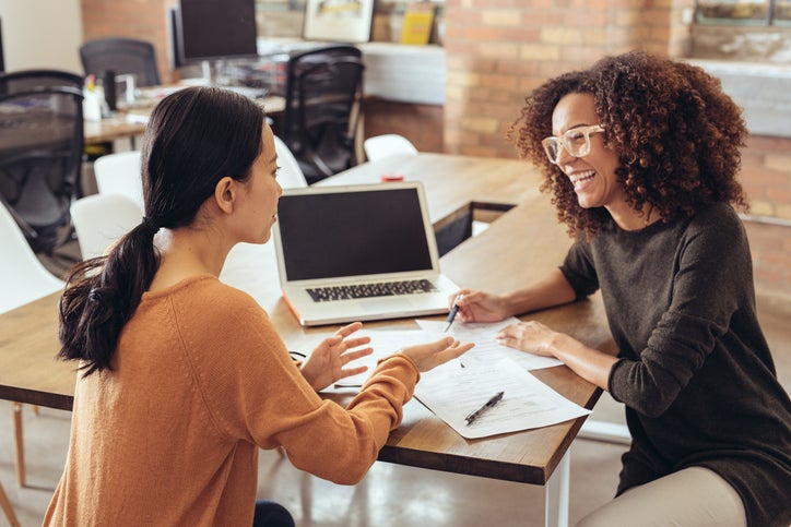 Two women sit at a desk with a pen, paper, and laptop, as one buys a business from the other