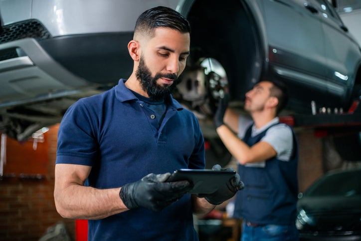 Supervisor at a car workshop checking tablet while an employee works at background on a car