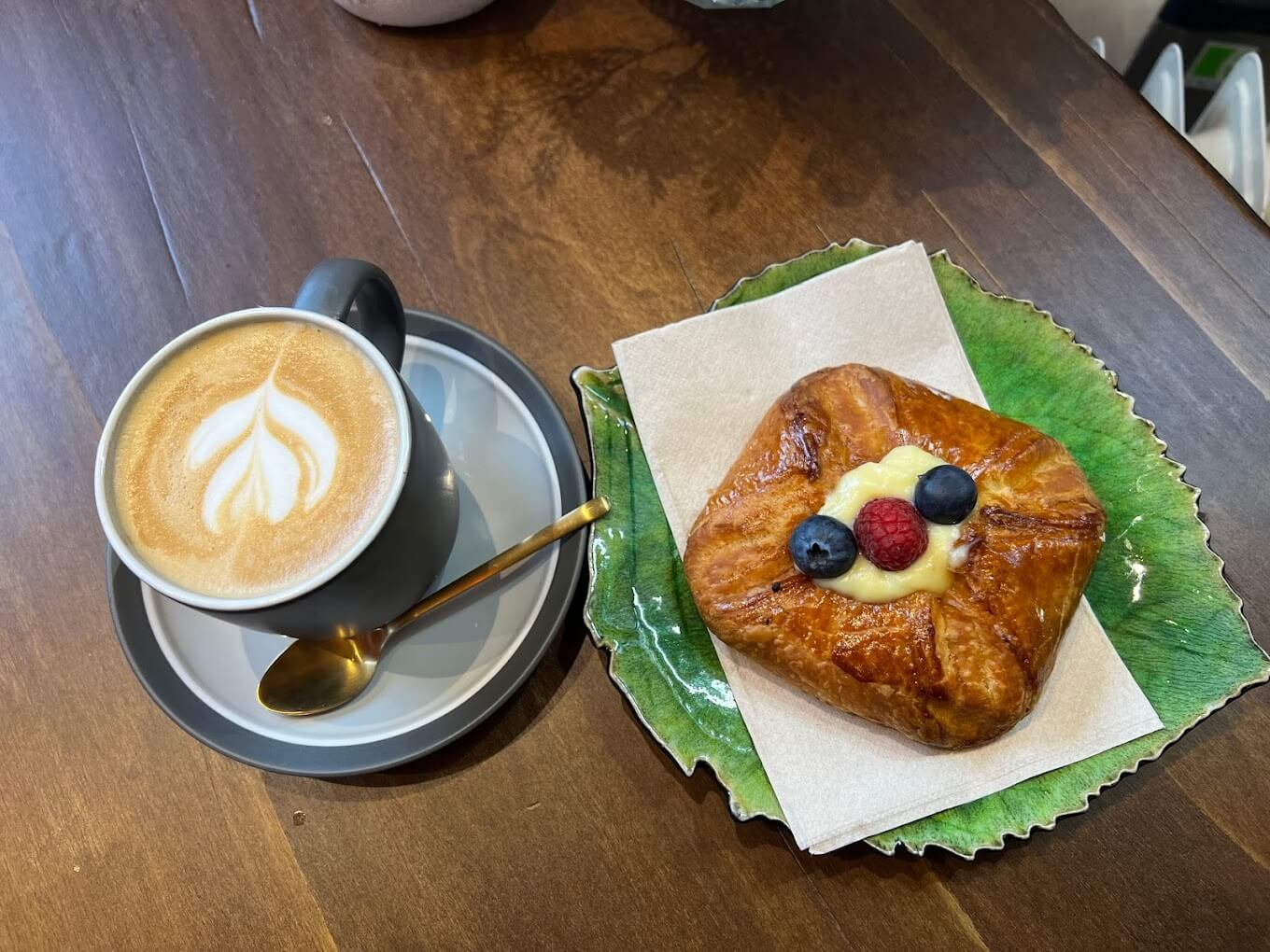 A cappuccino coffee in a cup on a saucer next to a fruit pasrty from the Cheerio Juice Bar