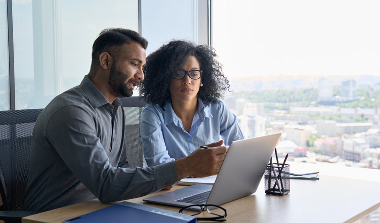 A business accountant speaks to a business owner who is using laptop sitting in modern office near panoramic window.