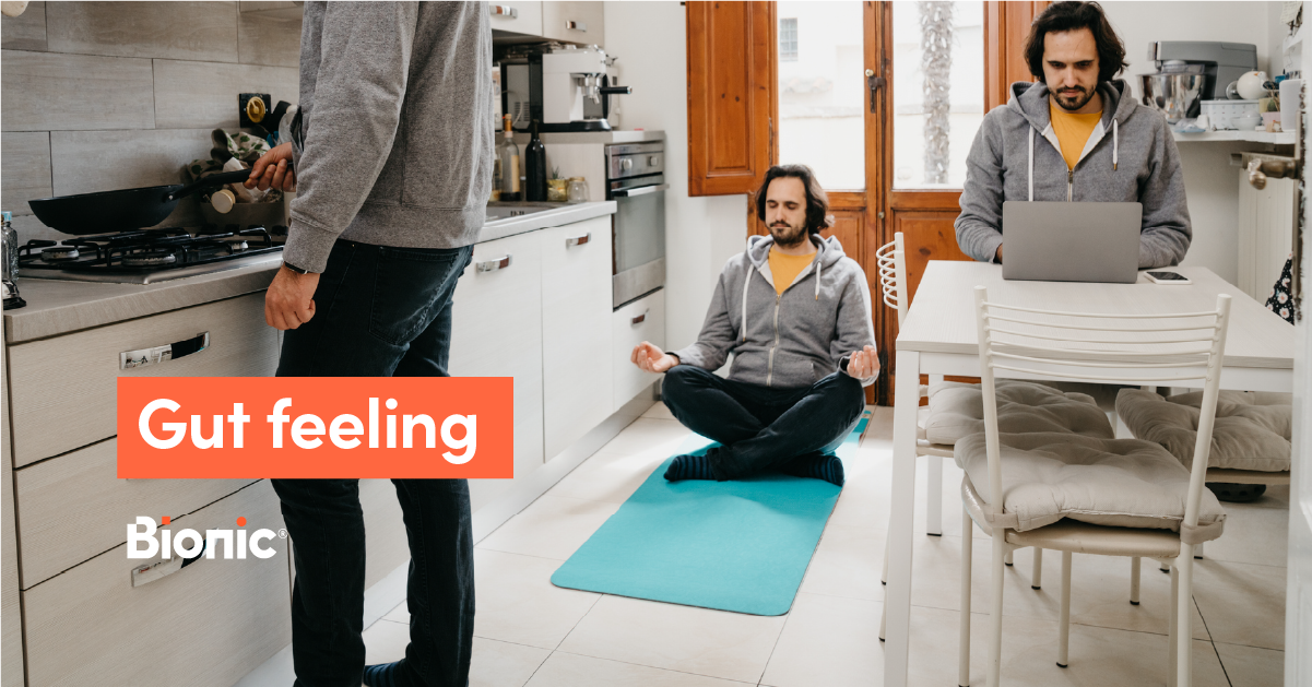 A man in a grey hoodie and blue trousers stands at a kitchen stove. He then sits cross legged on a turquoise yoga mat. He's finally shown working on a laptop while sitting at a white table.