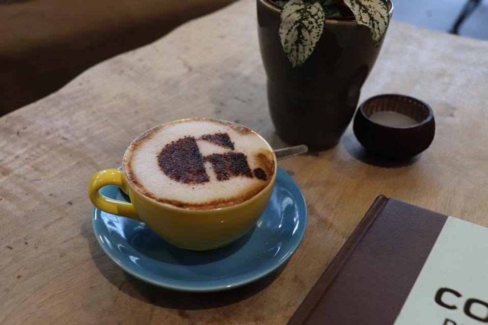 Yellow coffee cup on blue saucer on wooden table. Coffee cup has frothy cappuccino with Goupie logo on top in chocolate. 