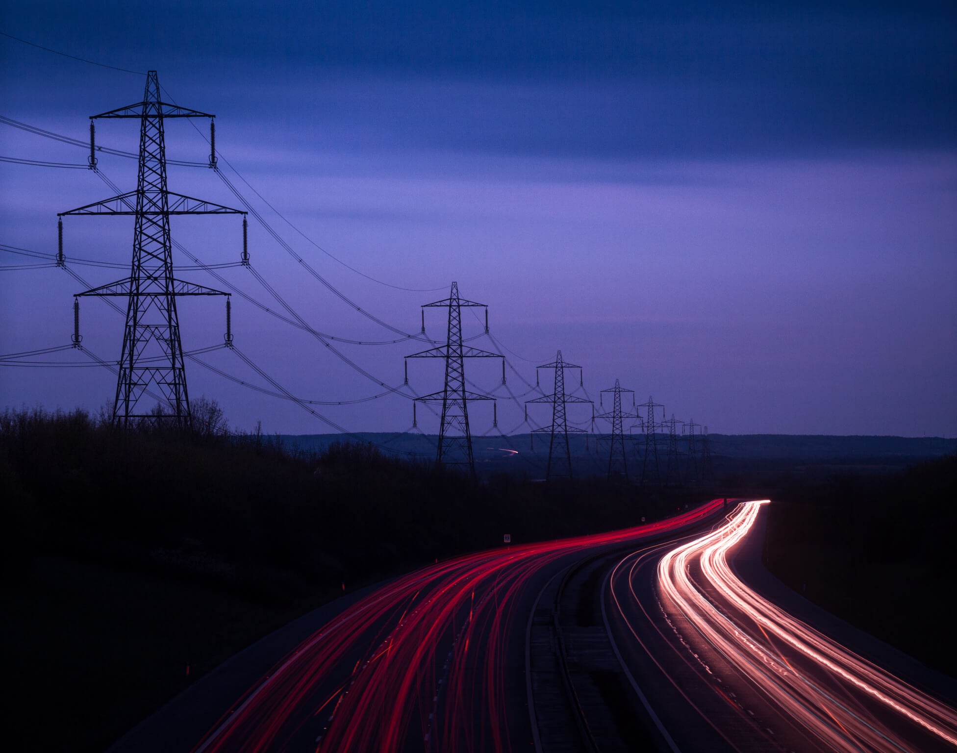 A series of electricity pylons silhouetted against a dusk sky. Cars pass on a motorway below.