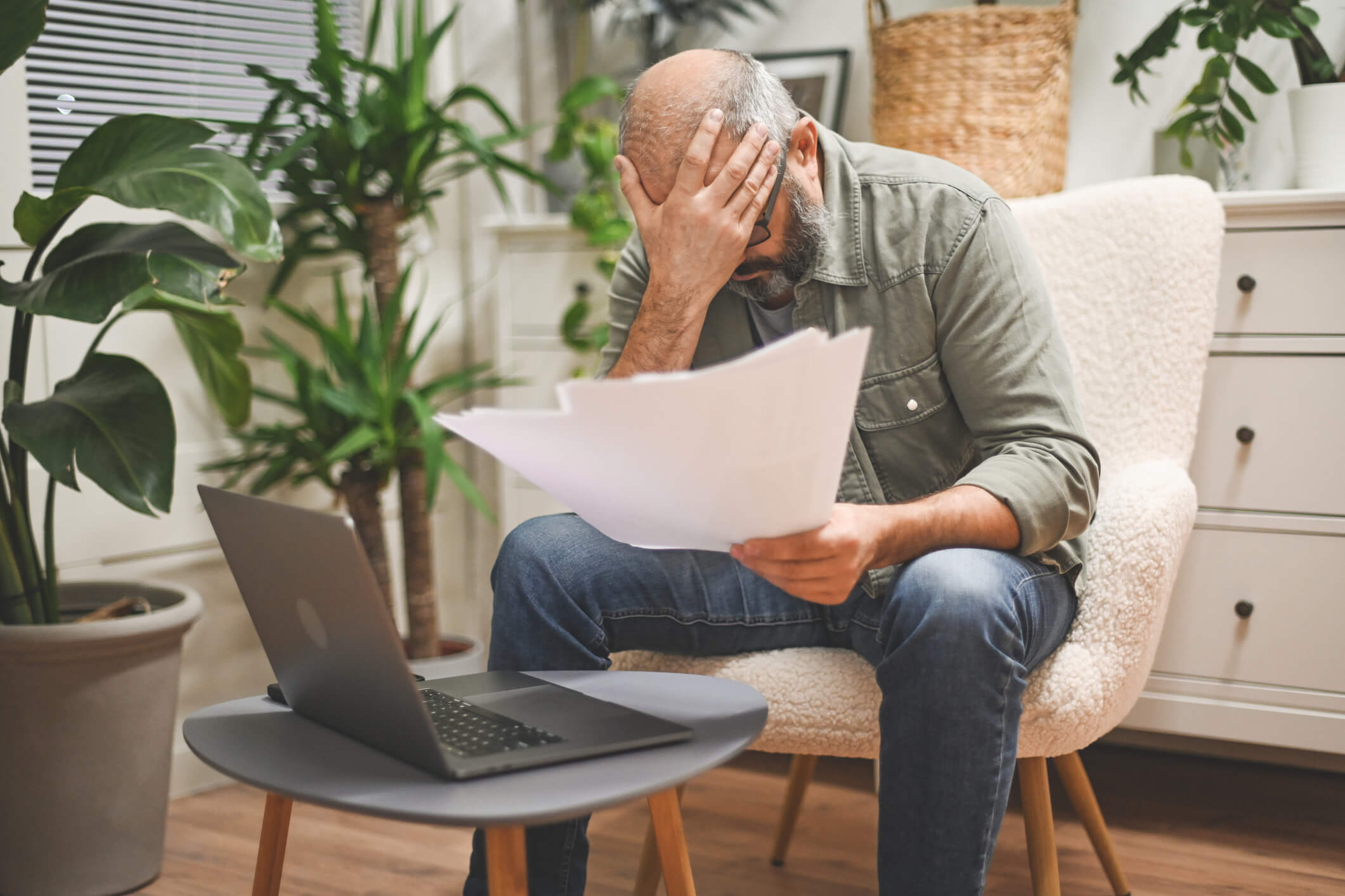 Man sat in a chair next to a laptop holding papers with his head in his hand