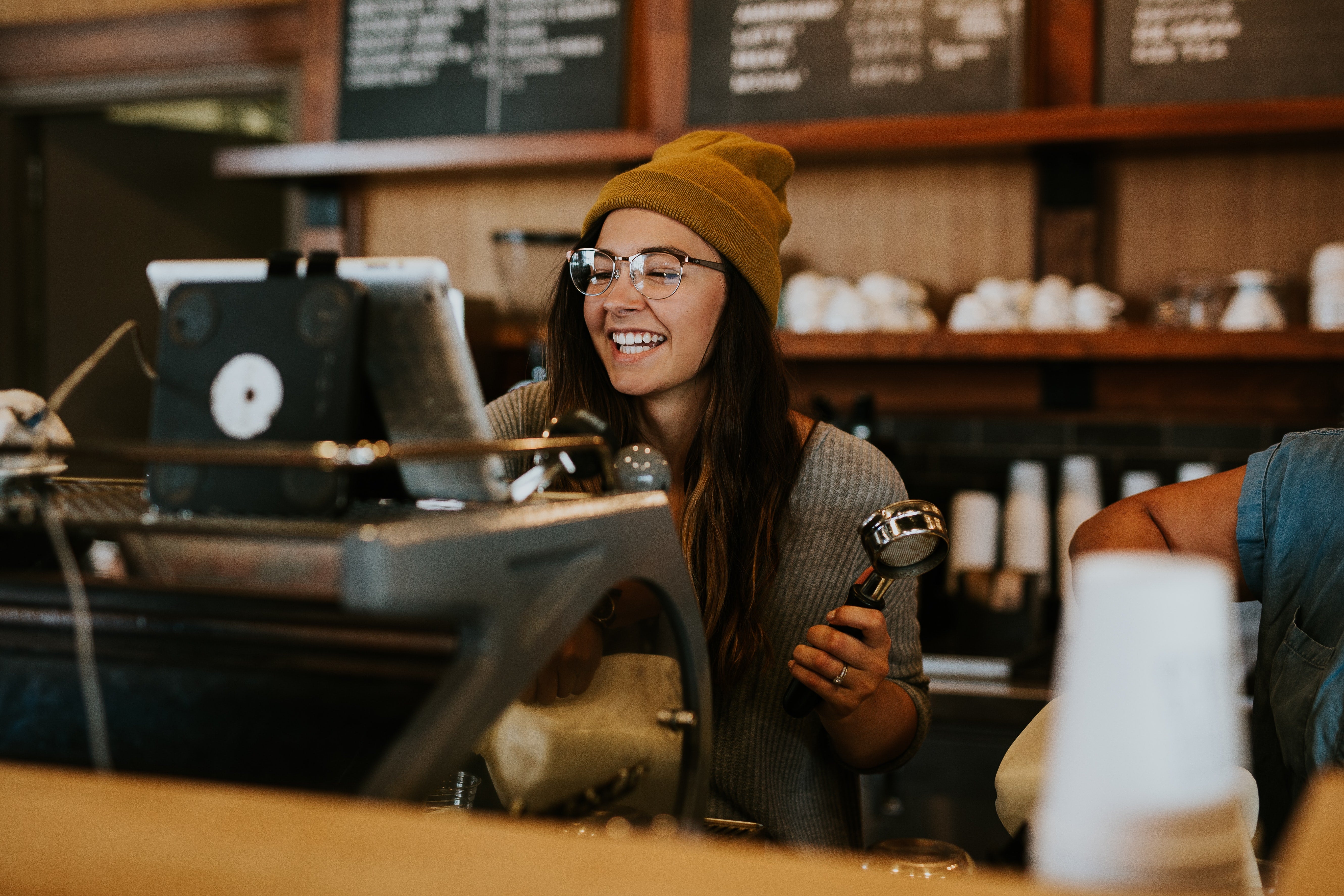 An independent coffee shop owner wearing glasses and a beanie hat smiles while serving a customer
