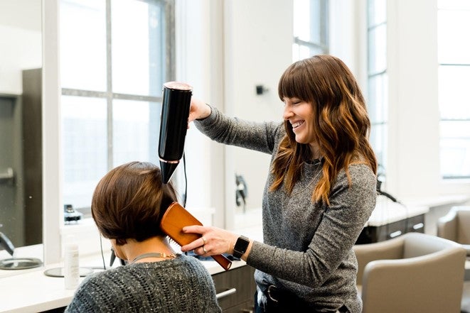 Female hairdresser with long brown hair brushes and blow dries a female client's short hair