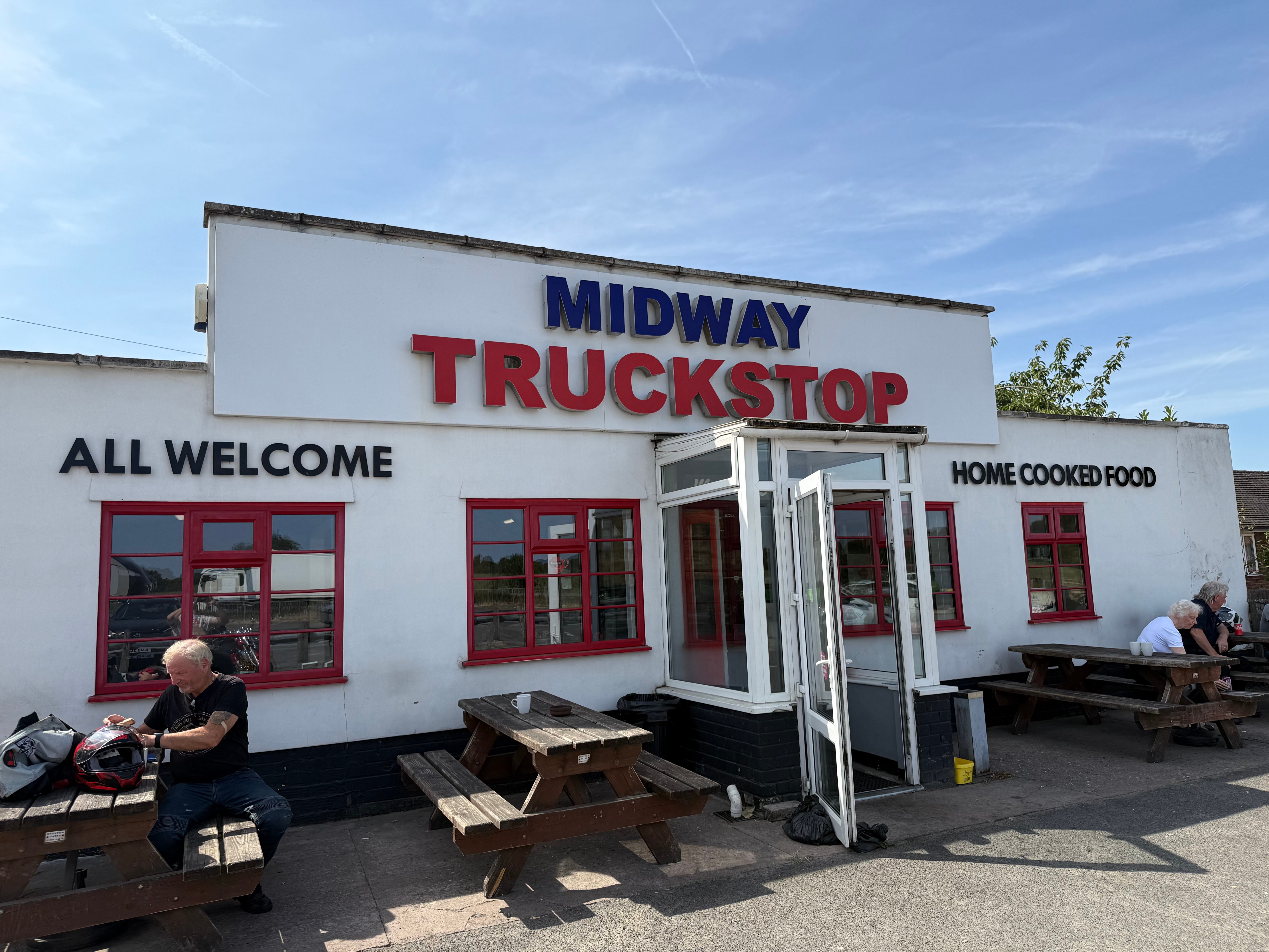 Midway Truckstop building with large red and blue signage, red-framed windows, and outdoor picnic benches where visitors are seated, beneath a clear blue sky.