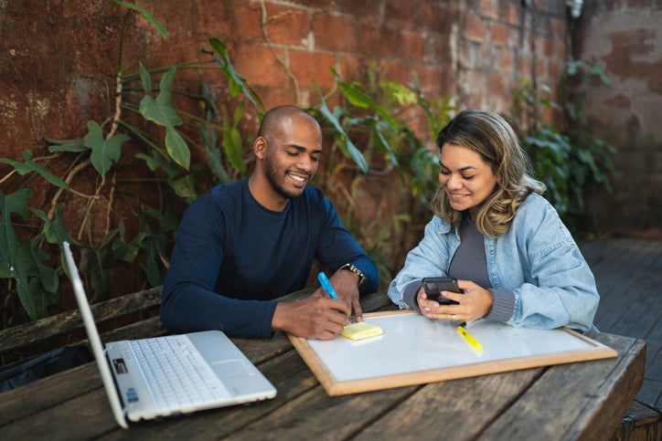 Two business owners sit at a desk with a white board and laptop to discuss a referral program