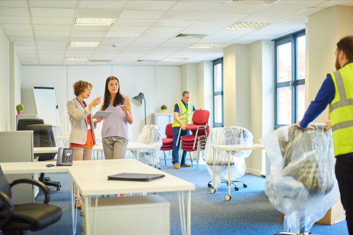 A new business venture is moving into some new offices. The owner is chatting about layout with her assistant as removal men bring in the office furniture.