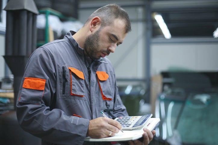 A mechanic stands in his workshop and uses a notepad and calculator to work out if he should get a tax refund