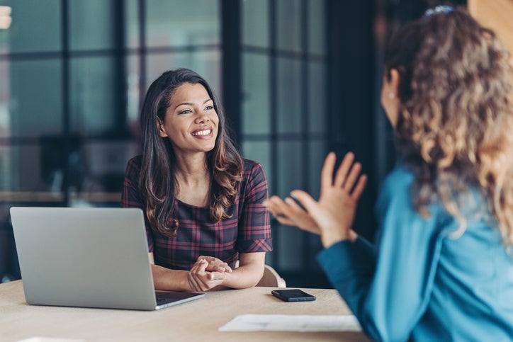 A woman in a checked dress sits at a laptop and interviews a candidate for a job