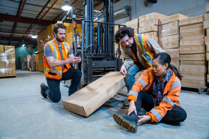 A worker sits down injured after being struck by a forklift. Employees help as she holds her ankle. Falls and collisions are covered by employers liability insurance