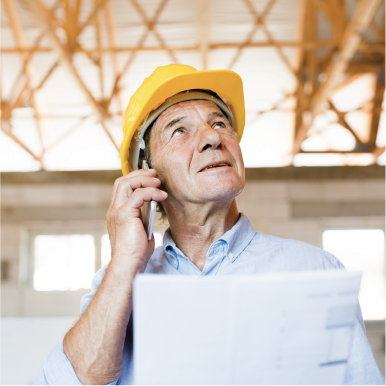 Man in blue shirt and yellow hard hat looks up while talking on mobile phone and reading energy bills.