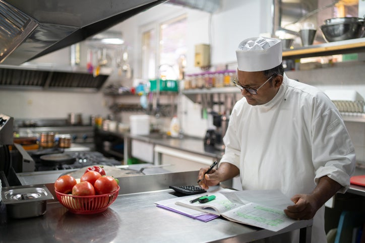 A restaurant owner stands in his kitchen. He has an energy bill and ledger in front of him as he works out energy costs