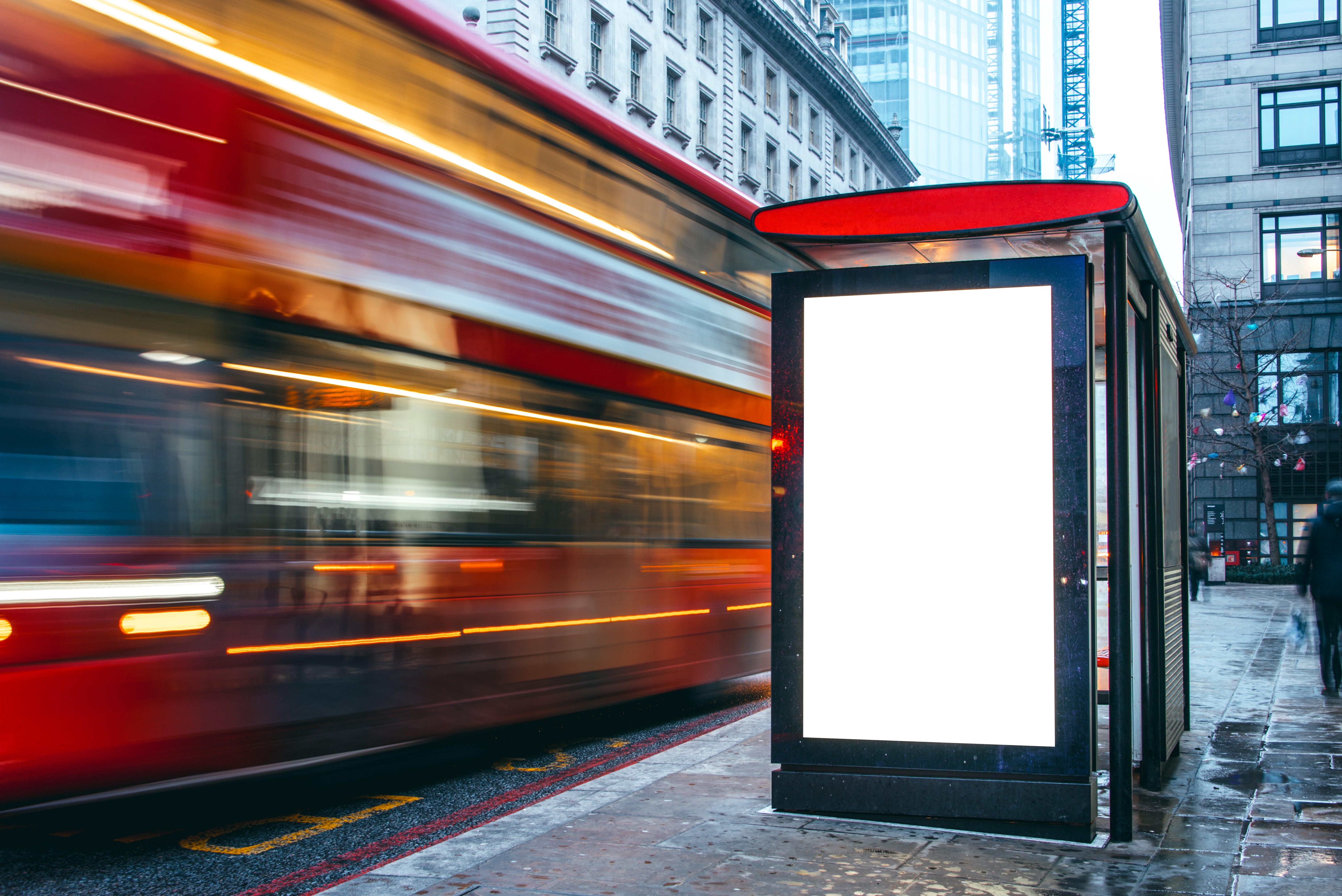 A blurred London bus drives past a bus stop with an blank advert poster on it
