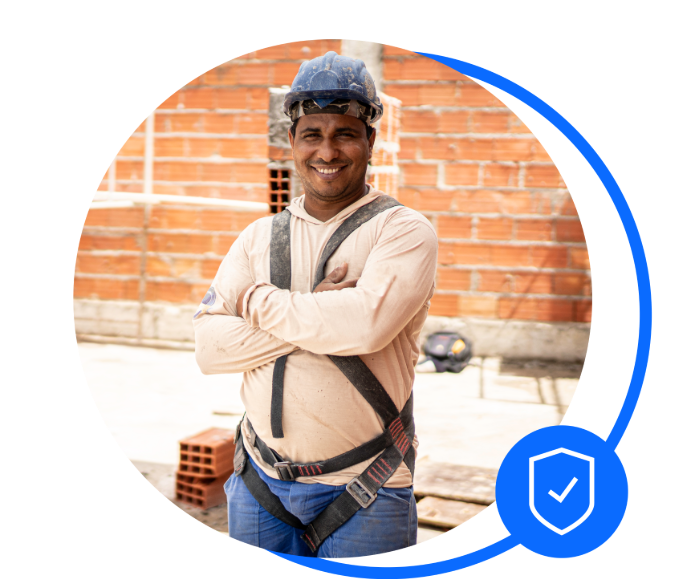 Male bricklayer stood cross armed in front of a brick wall and some red bricks. He's wearing a harness and a blue safety helmet. He's smiling at the camera