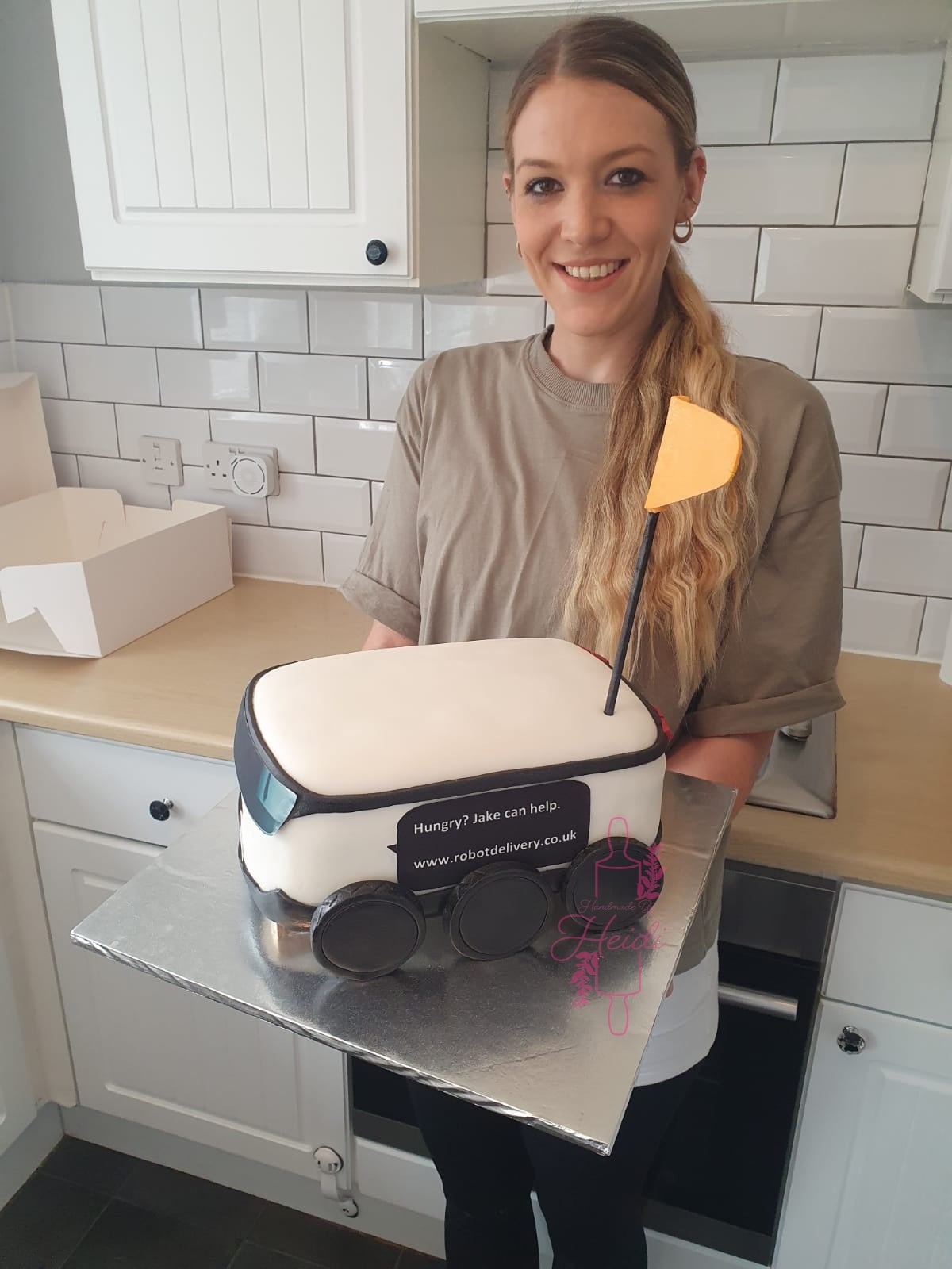 Baker Heidi Kay stands in her kitchen with a custom-made white go-kart cake with yellow flag