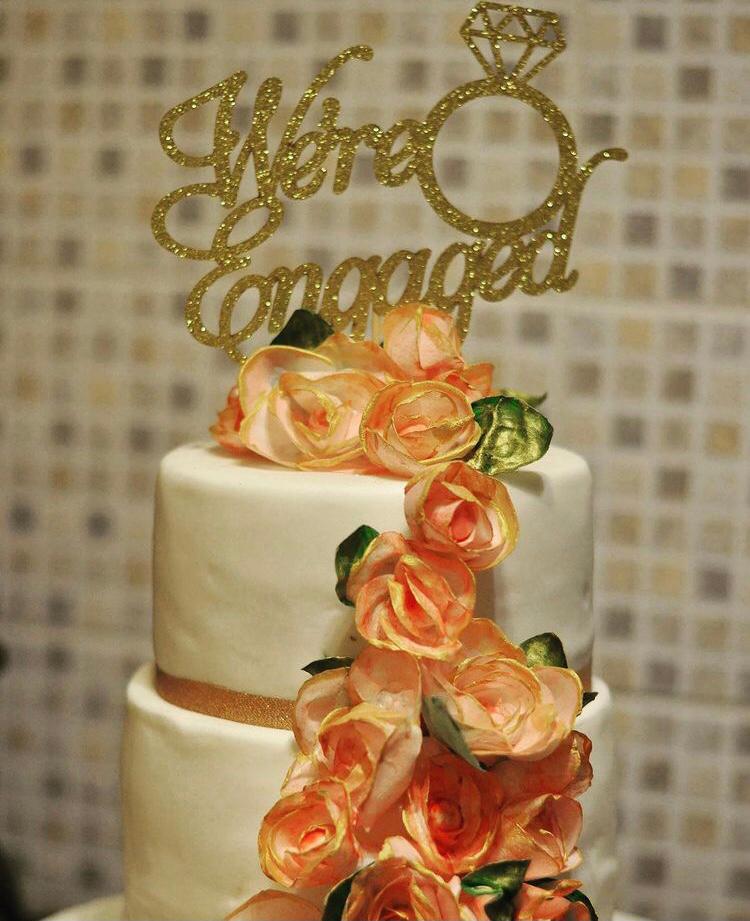 Multi-tiered engagement cake with white icing, handmade orange edible flowers and a gold 'We're Engaged' decoration