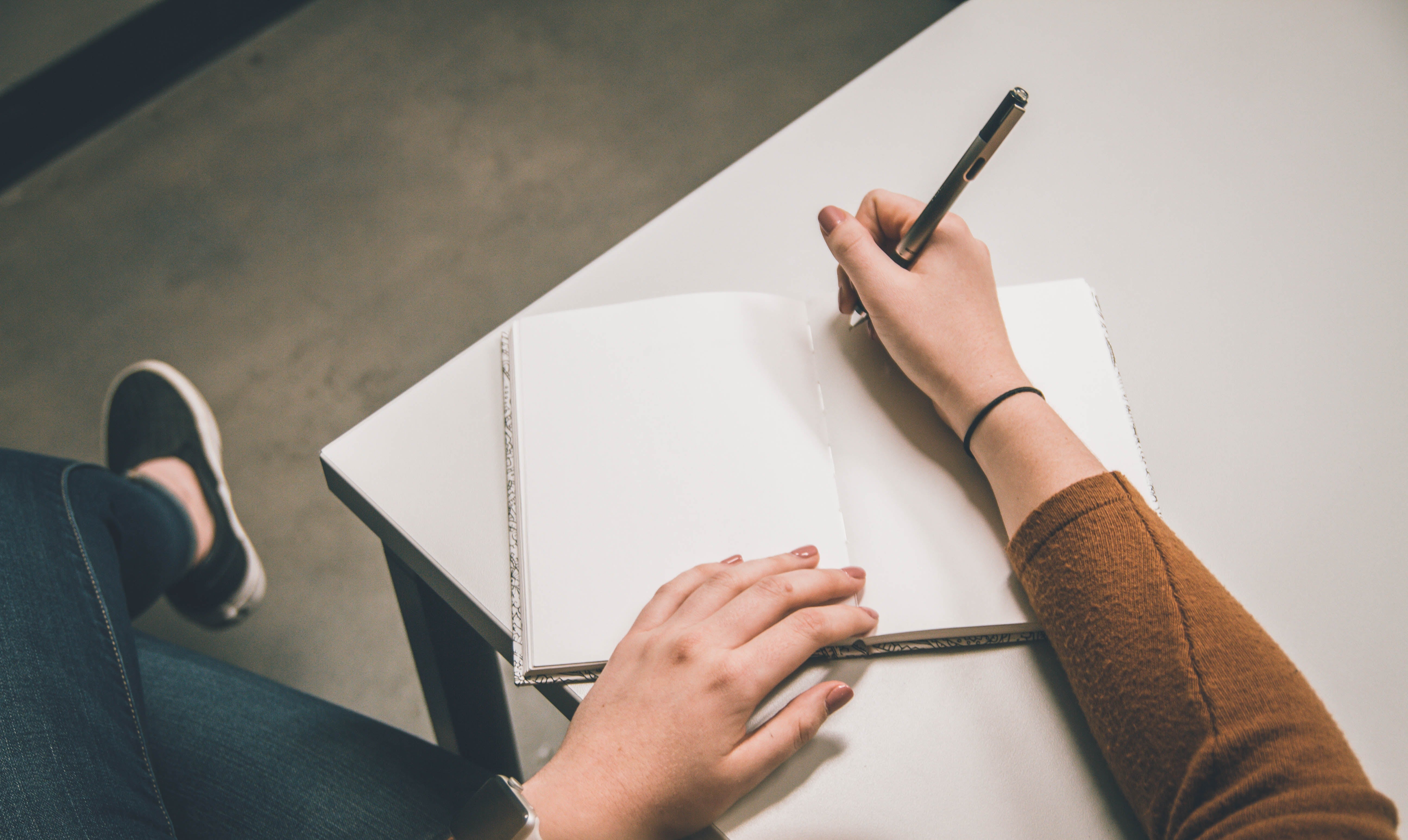 a woman in blue jeans, white pumps, and a brown top sits at a desk writing brand guidelines in a book with a black pen