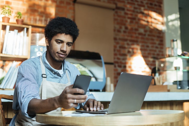 Small business owner wearing an apron sits at desk checking hs phone and laptop for equity finance options