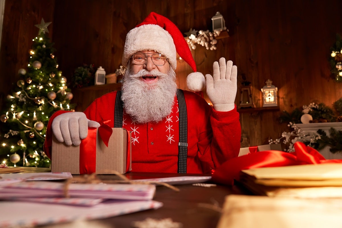 A man dressed as santa sat at a wooden table waving with a Christmas tree behind him