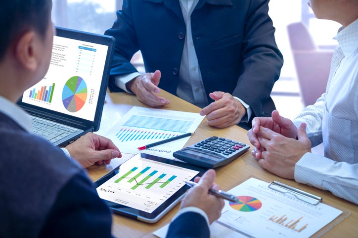 Three people in suits and shirts sit around a desk carrying out a business audit. A tablet touch screen is used to deeply reviewing a diagram or chart and financial reports for a return on investment or investment risk analysis or business performance.