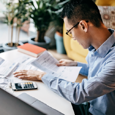 Commercial property owner wearing glasses and blue shirt looks at utility bills and works out costs on his smartphone calculator