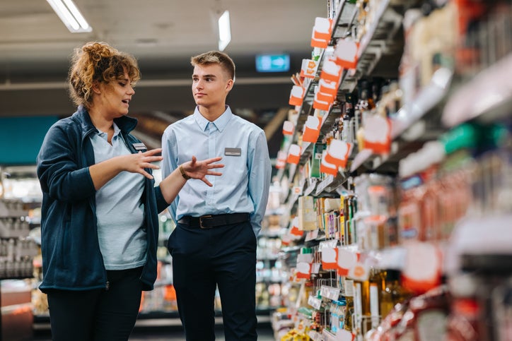 A shop owner in the aisle of her grocery shop is managing a staff member by talking him through what products are on sale.