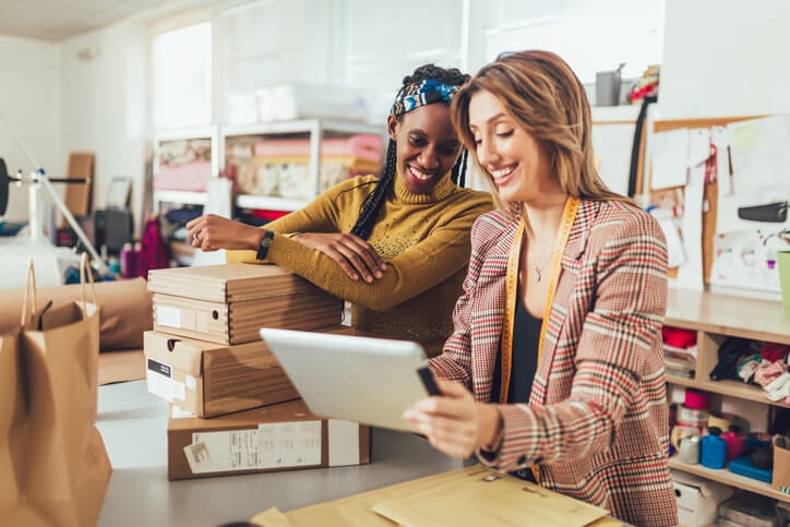 Two business owners checking their courier's delivery status on a a tablet while accepting new orders online and packing merchandise for customer.