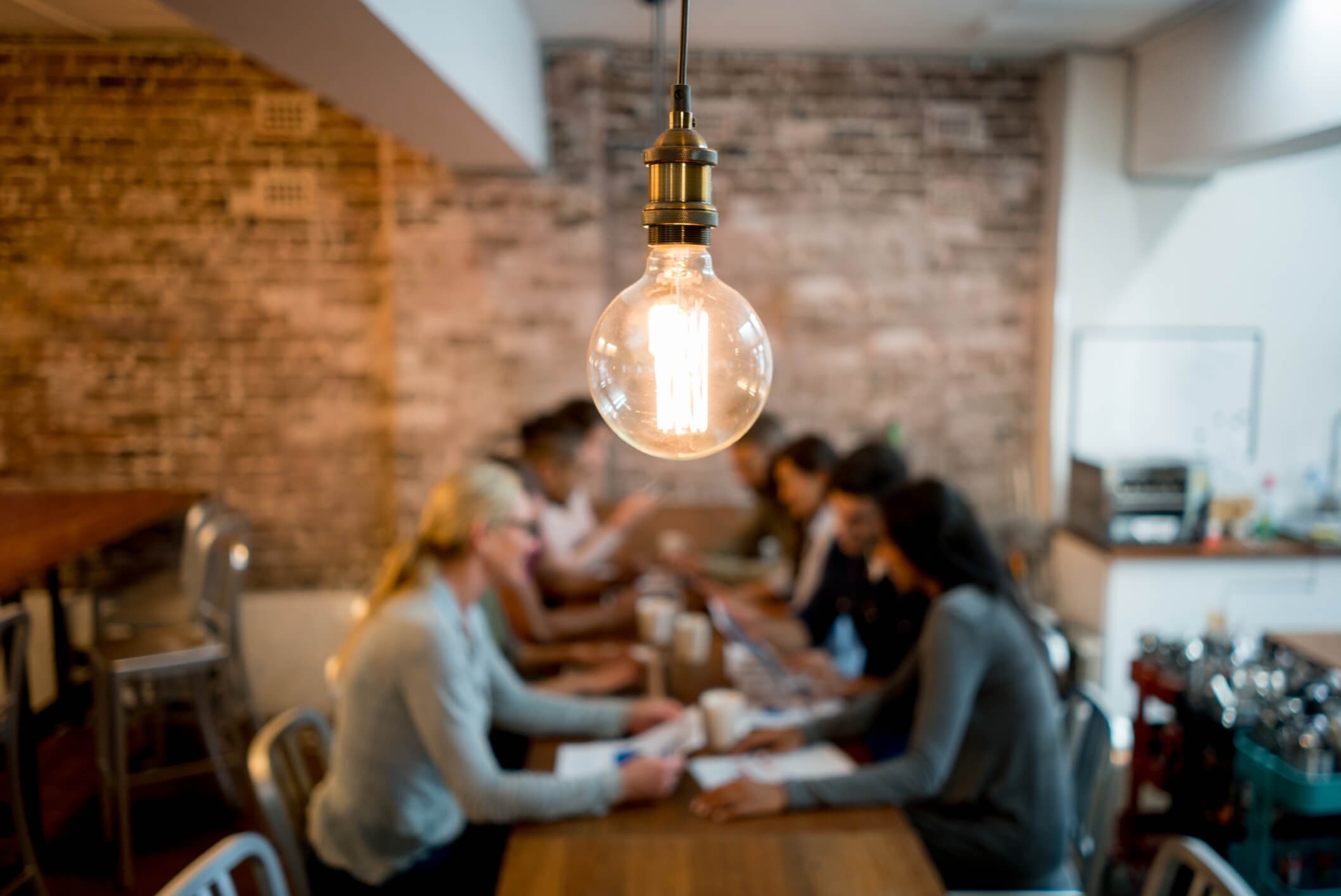 group of people sat at a long table in a coffee shop with coffees and paper with a lightbulb highlighted at the front