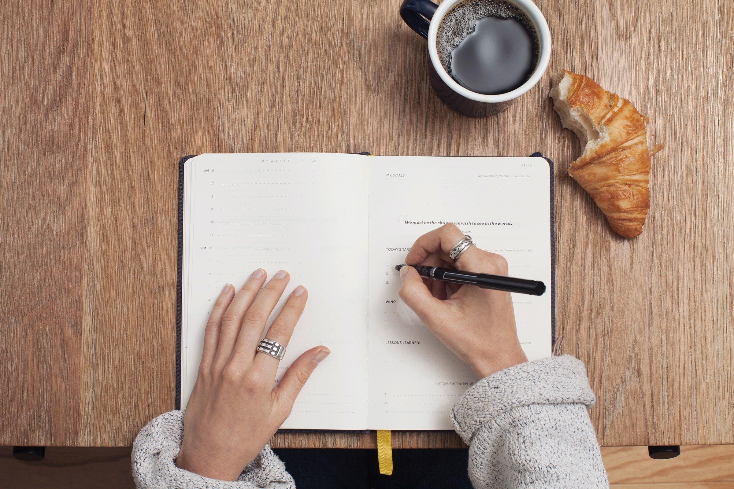 A woman in a grey sweater sits at a desk writing in a book with a black pen. A coffee and croissant is on the desk.