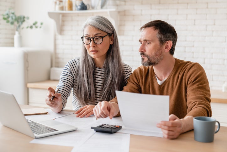 Two small business owners sit at a counter with a laptop, papers, and a calculator. They're planning a campaign and working out a marketing budget.